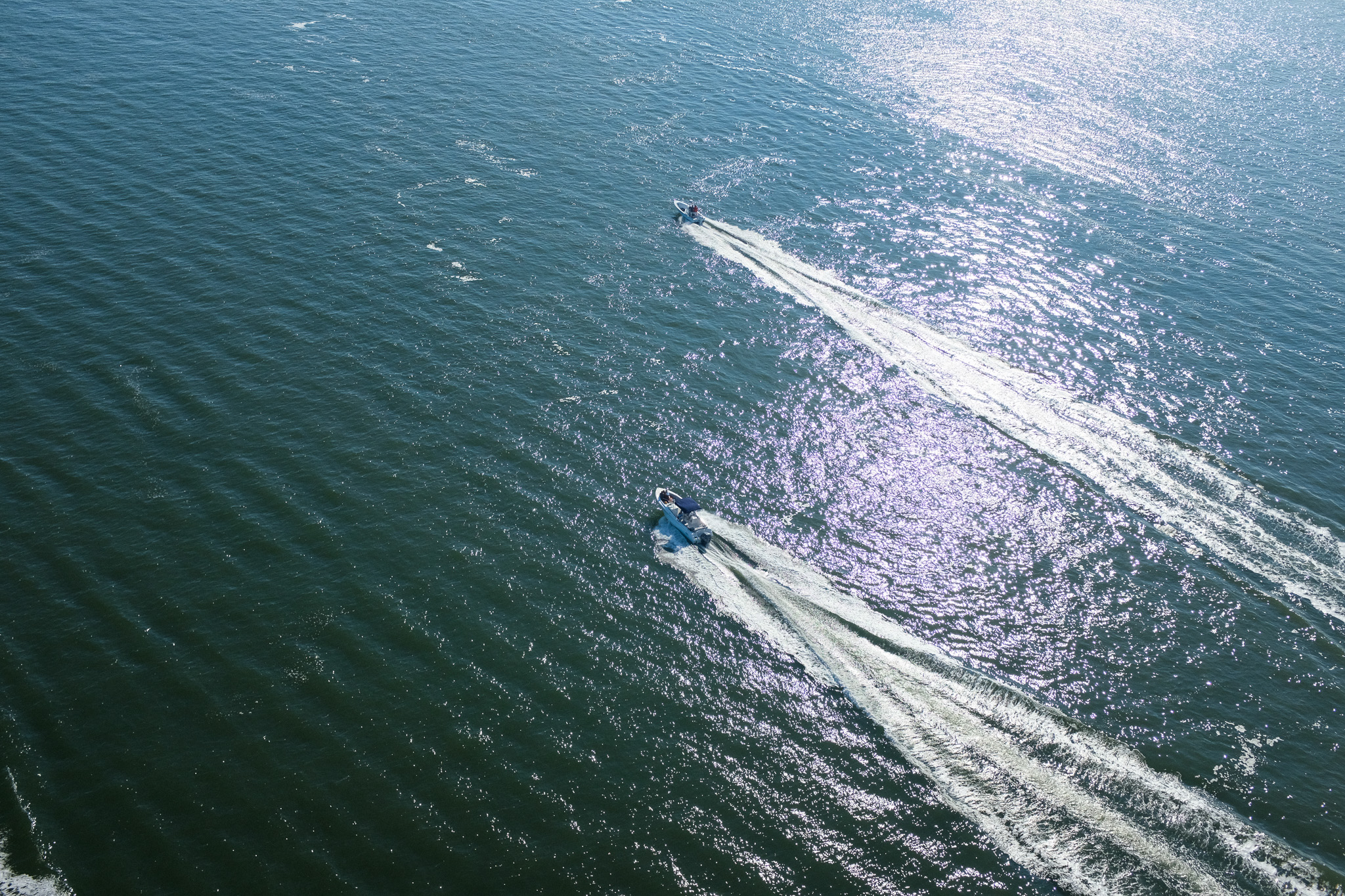 Aerial view of two small motorboats speeding across open blue water, leaving white wakes that sparkle in the sunlight.